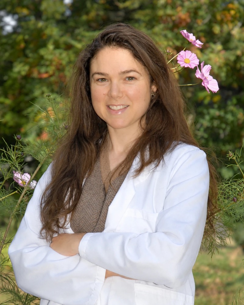 Belle Tenaglia smiling in a white coat, with pink cosmos flowers and greenery behind her